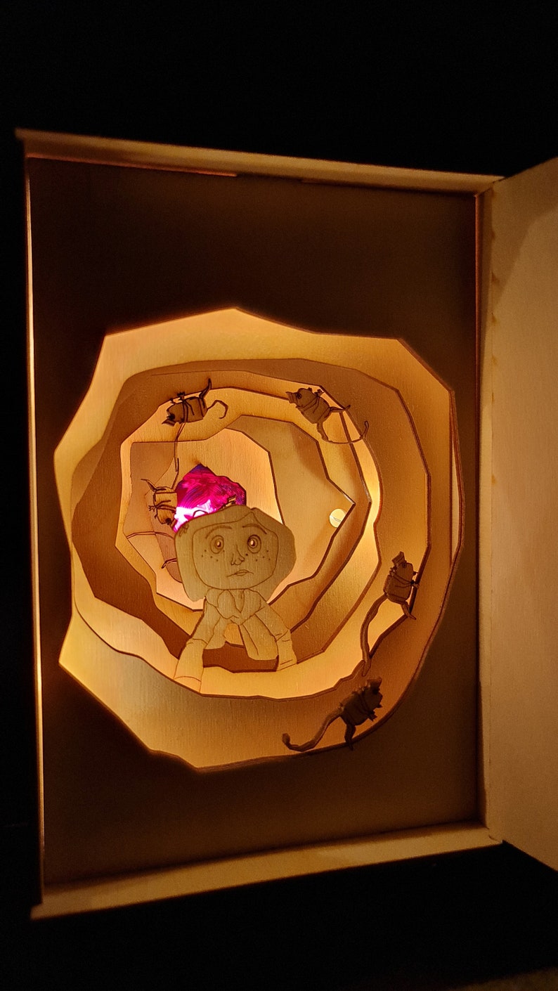 Pu&ograve; includere: Una scatola di legno con una luce all'interno, che crea un effetto 3D di una persona con un gioiello rosa sulla testa, circondata da piccole figure che salgono una scala a chiocciola.