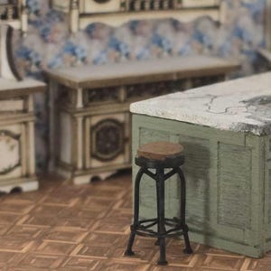 Puede incluir: Una escena de cocina en miniatura con un taburete de bar de metal negro y un asiento marrón. El taburete está frente a una encimera verde y blanca. El fondo incluye gabinetes y papel tapiz estampado, creando una estética vintage.