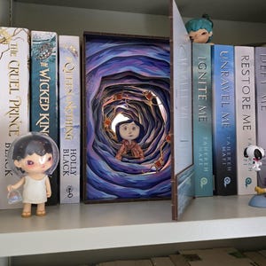 May include: A bookshelf with several books and decorative items. A 3D art piece depicting a scene from a movie is the focal point. A small figurine is on the left, and other figurines are on the right. The books have titles like "The Cruel Prince" and "Ignite Me."