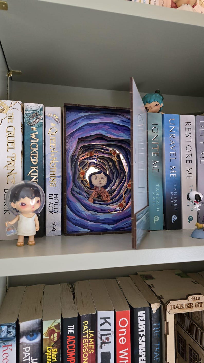 May include: A bookshelf with several books and decorative items. A 3D art piece depicting a scene from a movie is the focal point. A small figurine is on the left, and other figurines are on the right. The books have titles like "The Cruel Prince" and "Ignite Me."