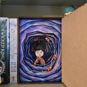 May include: A framed artwork depicting a young girl in a blue coat and hair, surrounded by a swirling tunnel of blue and purple hues. The artwork is displayed on a shelf with books.