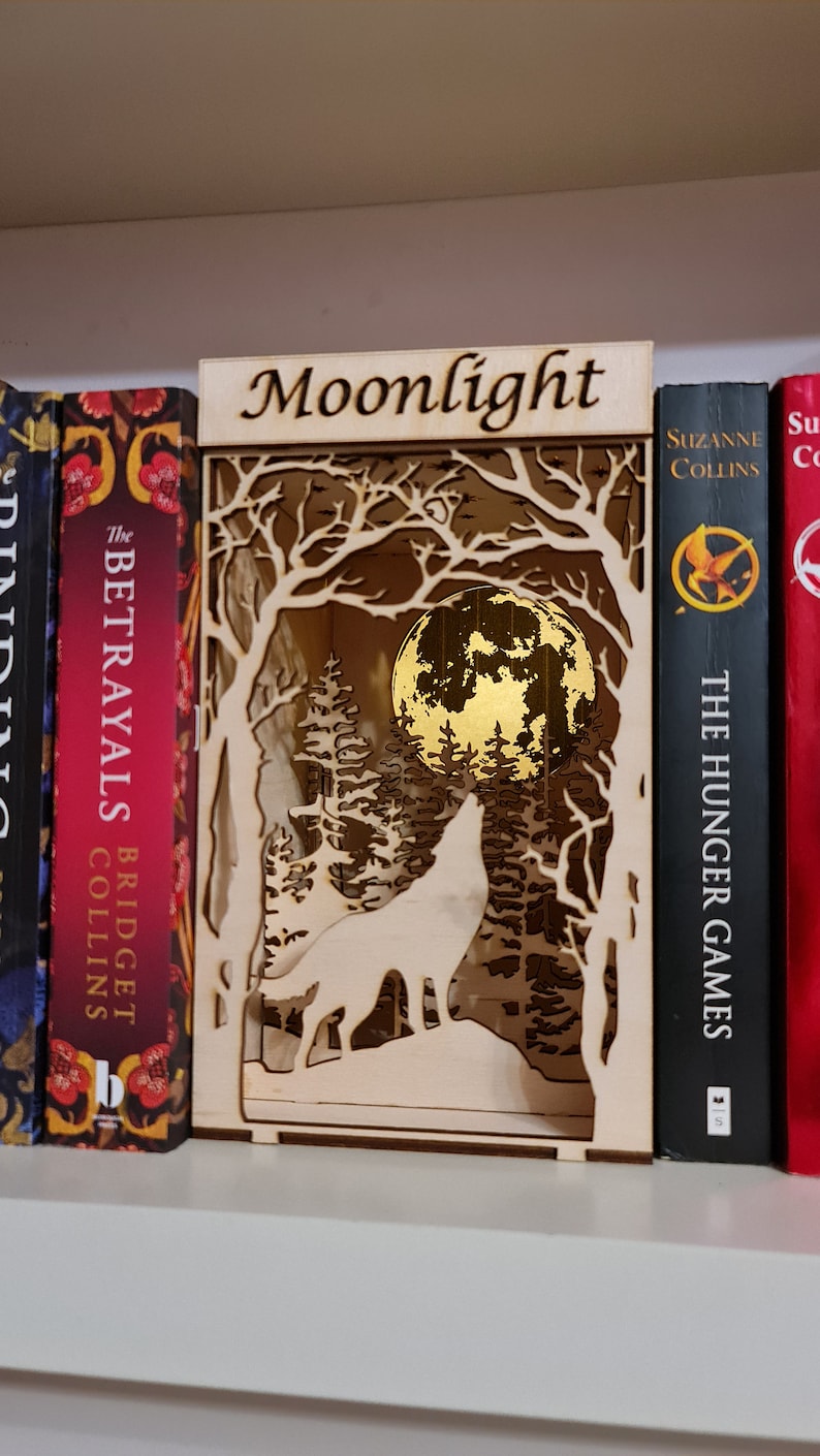 May include: A wooden bookend with a wolf howling at the moon in a forest scene. The bookend is labeled "Moonlight".