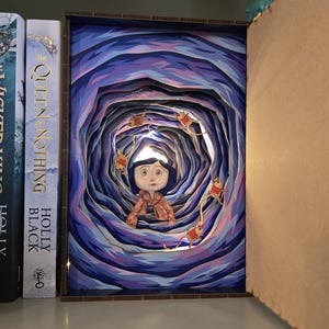 May include: A layered paper art piece depicting a character in a tunnel with a blue and purple color scheme. The artwork is illuminated with small lights, and the character is wearing a pink coat. The piece is displayed on a shelf next to books.