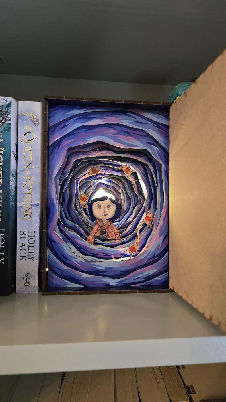 May include: A layered paper art piece depicting a character in a tunnel with a blue and purple color scheme. The artwork is illuminated with small lights, and the character is wearing a pink coat. The piece is displayed on a shelf next to books.