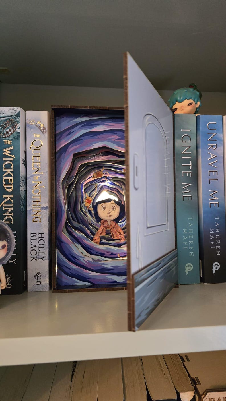May include: A layered paper art piece depicting a scene from a fantasy story, featuring a character standing in a tunnel. The artwork is set against a backdrop of books on a shelf. The color palette includes blues, purples, and browns.