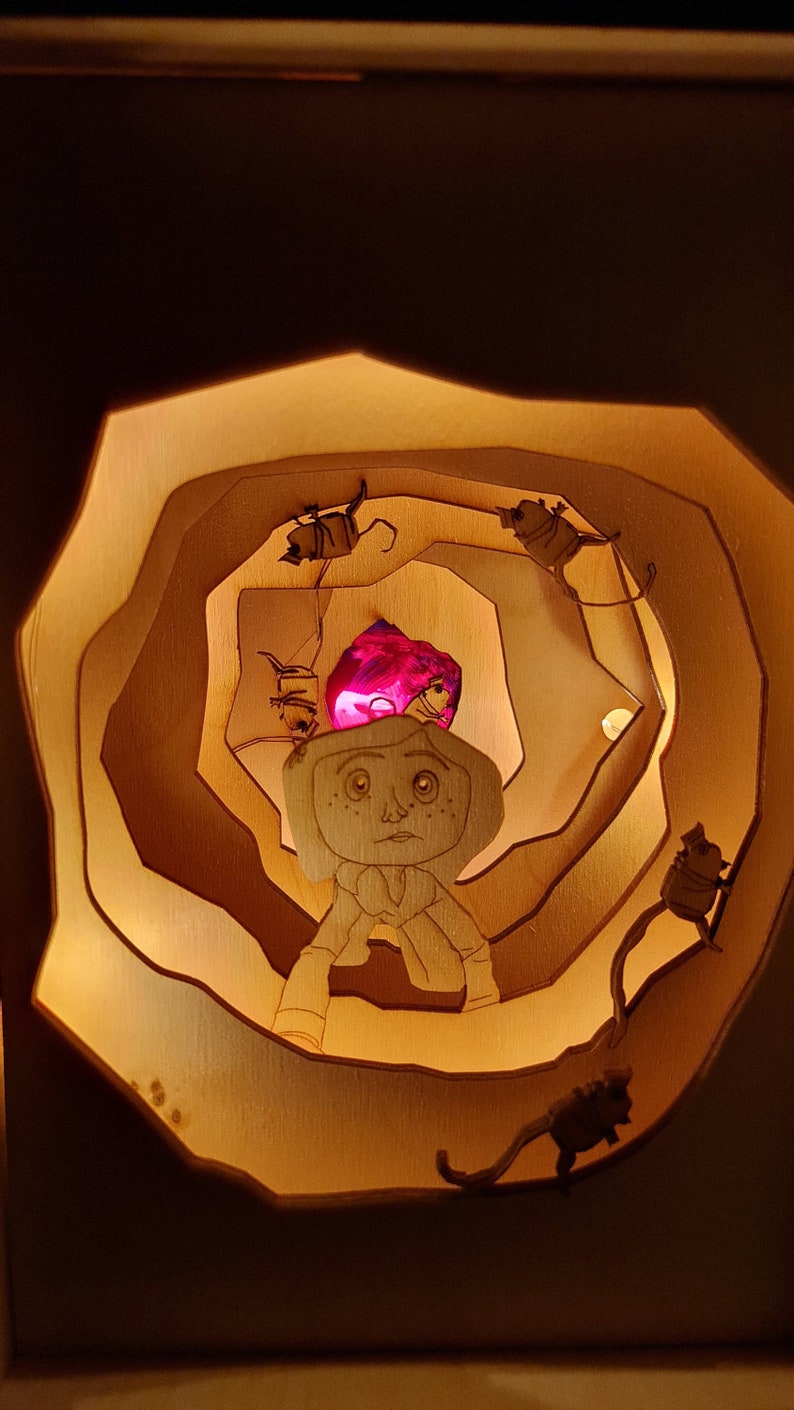 Pu&ograve; includere: Una scatola di legno illuminata con un design a strati. La scatola presenta un personaggio dei cartoni animati con una luce rosa che brilla dietro di lui. Il personaggio &egrave; circondato da piccole figure scure.