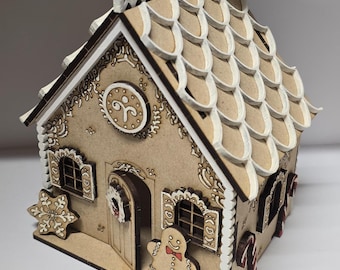 Può includere: Una dettagliata casa di pan di zenzero in legno con intricati disegni ritagliati. La casa presenta un tetto, finestre e porta rifiniti in bianco, con una figura di omino di pan di zenzero e accenti di bastoncini di zucchero. La combinazione di colori generale è marrone e bianco.