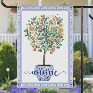 May include: A garden flag featuring a watercolor illustration of an orange tree in a blue and white pot. The flag has a blue border and the word "welcome" in script. The flag is displayed on a black metal stand.