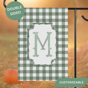 May include: A double-sided garden flag with a green and white checkered pattern. The flag features a light green letter "M" inside a decorative frame. The flag is displayed on a black metal stand, with an orange pumpkin in the foreground.