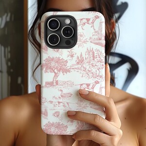 May include: A white iPhone case with a pink and white toile pattern featuring hunting dogs, trees, and a small building. The case has a black camera lens cover.