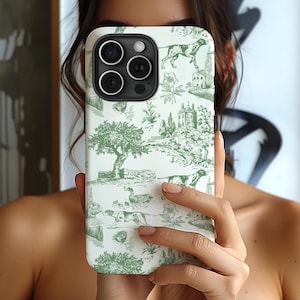 May include: A white iPhone case with a green and white toile pattern featuring hunting dogs, birds, and trees. The case has a camera lens cutout and a raised lip around the edges.