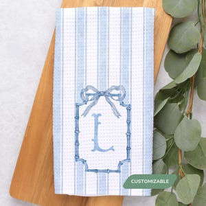 May include: A white and blue striped kitchen towel with a monogrammed letter "L" in a bamboo-style frame. The towel has a bow at the top of the frame and is labeled "Customizable".