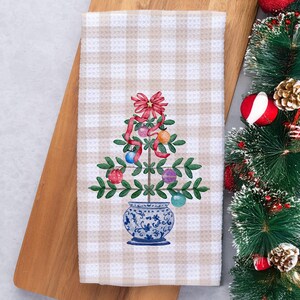 May include: A beige and white checkered kitchen towel featuring a watercolor Christmas tree design. The tree is decorated with colorful ornaments and a red bow. The tree sits in a blue and white vase. The towel is on a wooden cutting board.