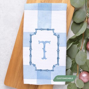 May include: A white and blue checkered kitchen towel with a blue monogram letter "T" inside a bamboo-style frame. The towel is on a wooden cutting board, with eucalyptus and ornaments in the background. The word "CUSTOMIZABLE" is in the bottom right corner.