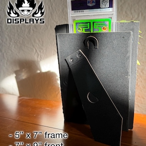 May include: A black display frame with a flexible kickstand, holding a PSA graded trading card in a clear plastic case. The frame features a 5 inch by 7 inch interior and a 7 inch by 9 inch front. The Fire Type Displays logo is visible in the corner.