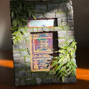 May include: A rectangular display frame with a textured gray stone brick finish and green moss accents. The frame features a top slot holding a PSA graded 2000 Pokemon Ancient Mew card and a larger central window displaying a card with the text Card not included.