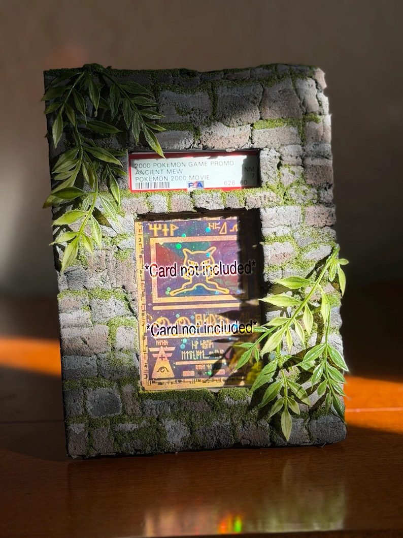 May include: A rectangular display frame with a textured gray stone brick finish and green moss accents. The frame features a top slot holding a PSA graded 2000 Pokemon Ancient Mew card and a larger central window displaying a card with the text Card not included.