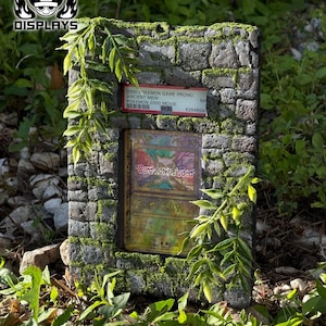 May include: A rectangular stone-textured display frame featuring green moss accents and artificial leafy vines. The frame holds a PSA-graded 2000 Pokemon Game Promo Ancient Mew card and includes a central placeholder text reading Card not included.