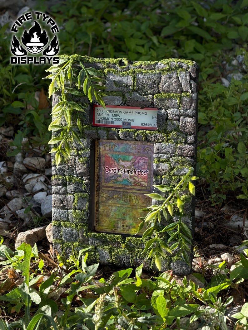 May include: A rectangular stone-textured display frame featuring green moss accents and artificial leafy vines. The frame holds a PSA-graded 2000 Pokemon Game Promo Ancient Mew card and includes a central placeholder text reading Card not included.