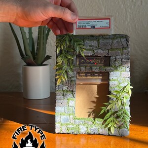 May include: A rectangular display stand designed to look like a weathered stone wall with green moss accents and artificial leafy vines. The structure features a slot at the top for a trading card slab and a central rectangular opening for display.