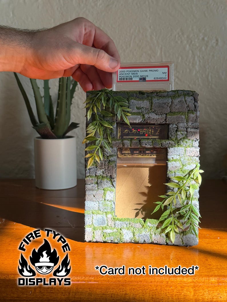 May include: A rectangular display stand designed to look like a weathered stone wall with green moss accents and artificial leafy vines. The structure features a slot at the top for a trading card slab and a central rectangular opening for display.
