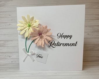 May include: A white card with a pink and yellow paper flower bouquet, a white tag with the name "Toni" and the text "Happy Retirement".
