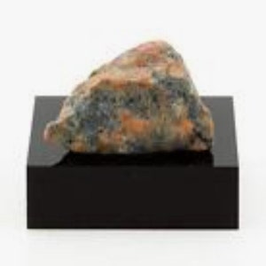 Puede incluir: Una pequeña roca de forma irregular con una mezcla de colores naranja, azul y gris. La roca se muestra sobre una base rectangular negra.