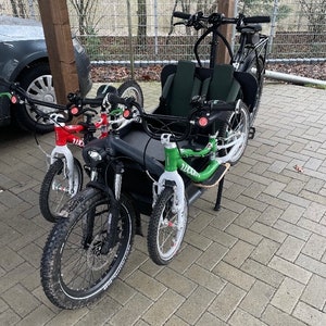 Könnte beinhalten: Drei schwarze Fahrräder mit weißen Reifen und roten Lenkern. Die Fahrräder stehen auf einer grauen Pflasterfläche. Das Fahrrad hinten hat einen grünen Sattel und einen schwarzen Rahmen.