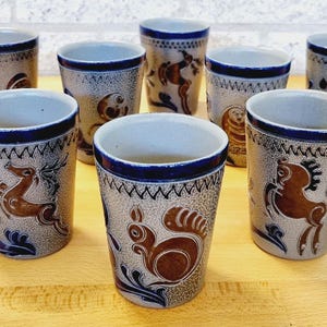 Op de afbeelding: Een set van zeven keramische bekers met een gespikkelde grijze afwerking en handgeschilderde bruine diermotieven, waaronder eekhoorns en herten, geaccentueerd door donkerblauwe randen en decoratieve zigzagpatronen langs de bovenste randen.