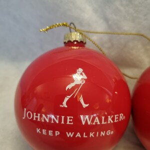 May include: Red Christmas ornament with the Johnnie Walker logo and the words "JOHNNIE WALKER KEEP WALKING" in white. The ornament has a gold string for hanging.