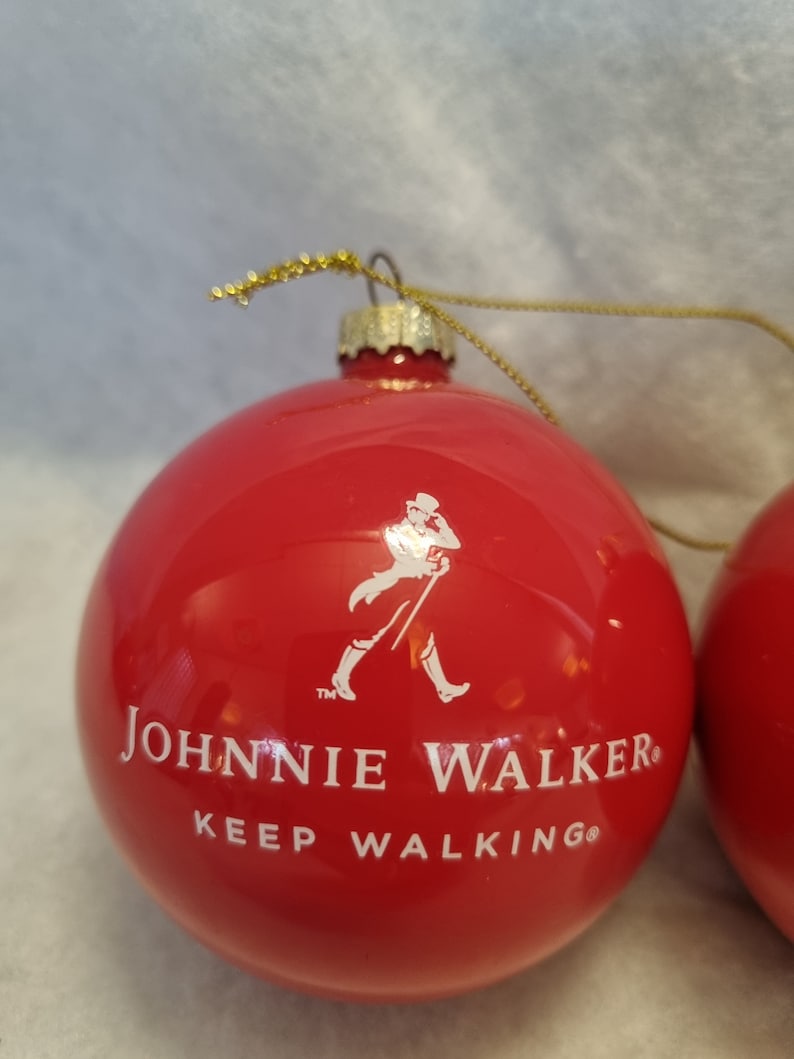 May include: Red Christmas ornament with the Johnnie Walker logo and the words "JOHNNIE WALKER KEEP WALKING" in white. The ornament has a gold string for hanging.