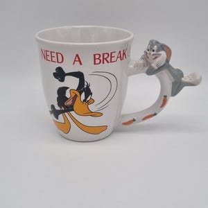 Op de afbeelding: Witte keramische mok met een cartoon afbeelding van Daffy Duck aan de voorkant. De mok heeft een handvat in de vorm van Bugs Bunny met wortel accenten. De tekst "NEED A BREAK" is in rood op de mok gedrukt.