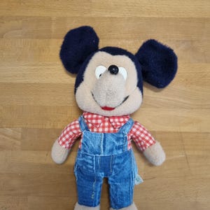 May include: A plush Mickey Mouse doll with navy blue ears, a tan face, and a red and white checkered shirt. The doll is wearing blue denim overalls and has tan feet.