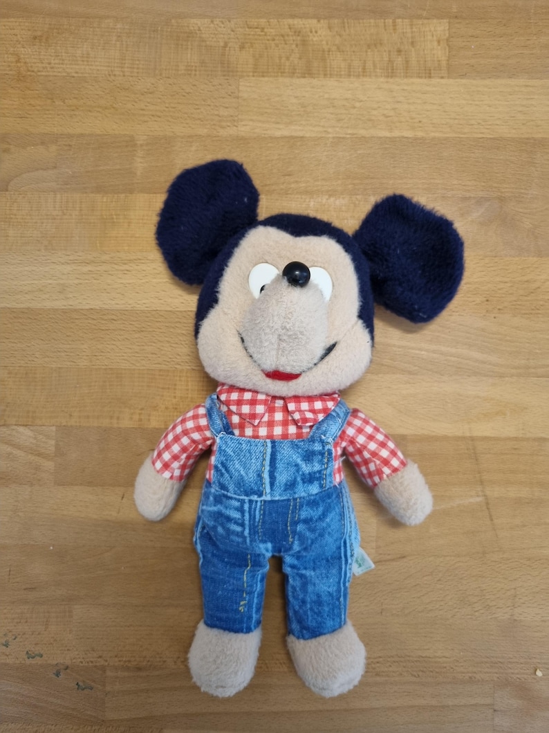 May include: A plush Mickey Mouse doll with navy blue ears, a tan face, and a red and white checkered shirt. The doll is wearing blue denim overalls and has tan feet.