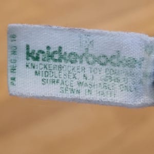 May include: A light blue tag with green text. The tag reads "knickerbocker" and includes the text "KNICKERBOCKER TOY COMPANY MIDDLESEX, N.J." and "SURFACE WASHABLE ONLY SEWN IN HAITI."