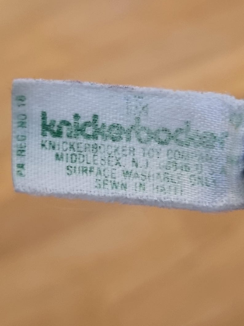May include: A light blue tag with green text. The tag reads "knickerbocker" and includes the text "KNICKERBOCKER TOY COMPANY MIDDLESEX, N.J." and "SURFACE WASHABLE ONLY SEWN IN HAITI."