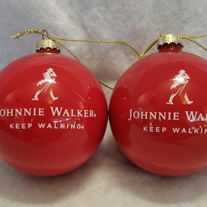 May include: Two red Christmas ornaments with the Johnnie Walker logo and the words "KEEP WALKING" in white. Each ornament has a gold-colored string for hanging.