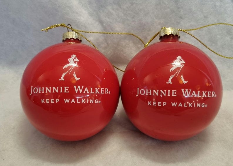 May include: Two red Christmas ornaments with the Johnnie Walker logo and the words "KEEP WALKING" in white. Each ornament has a gold-colored string for hanging.