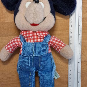 May include: A plush Mickey Mouse doll wearing blue denim overalls and a red and white checkered shirt. The doll has black ears, a black nose, and a smiling red mouth. A ruler is placed next to the doll.