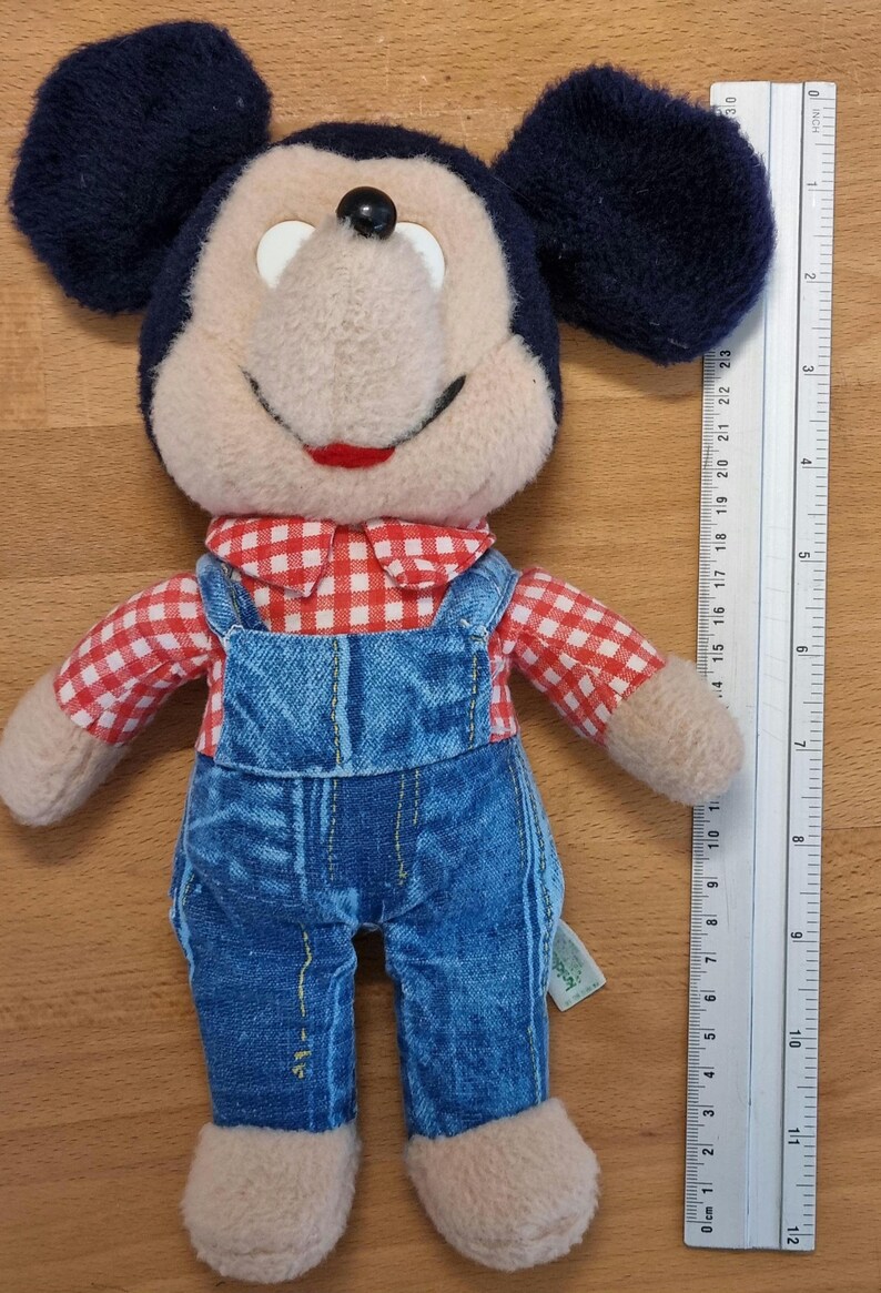May include: A plush Mickey Mouse doll wearing blue denim overalls and a red and white checkered shirt. The doll has black ears, a black nose, and a smiling red mouth. A ruler is placed next to the doll.