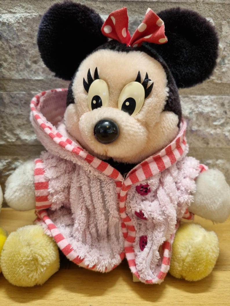 May include: A plush toy of Mickey Mouse wearing a pink and red striped robe. The toy has black ears, a red bow with white polka dots, and large black eyes.