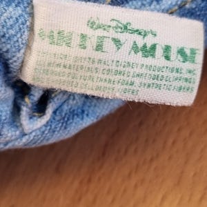 May include: A close-up of a white tag with green text that reads "Walt Disney's MICKEY MOUSE". The tag is attached to a blue denim fabric. The tag also includes additional text about the materials used.