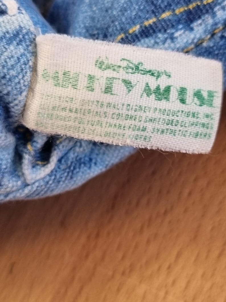 May include: A close-up of a white tag with green text that reads "Walt Disney's MICKEY MOUSE". The tag is attached to a blue denim fabric. The tag also includes additional text about the materials used.