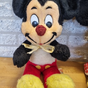 May include: Vintage plush Mickey Mouse toy with black ears, a red and yellow outfit, and a yellow bow tie. The toy has a smiling face with black eyes and a red nose.