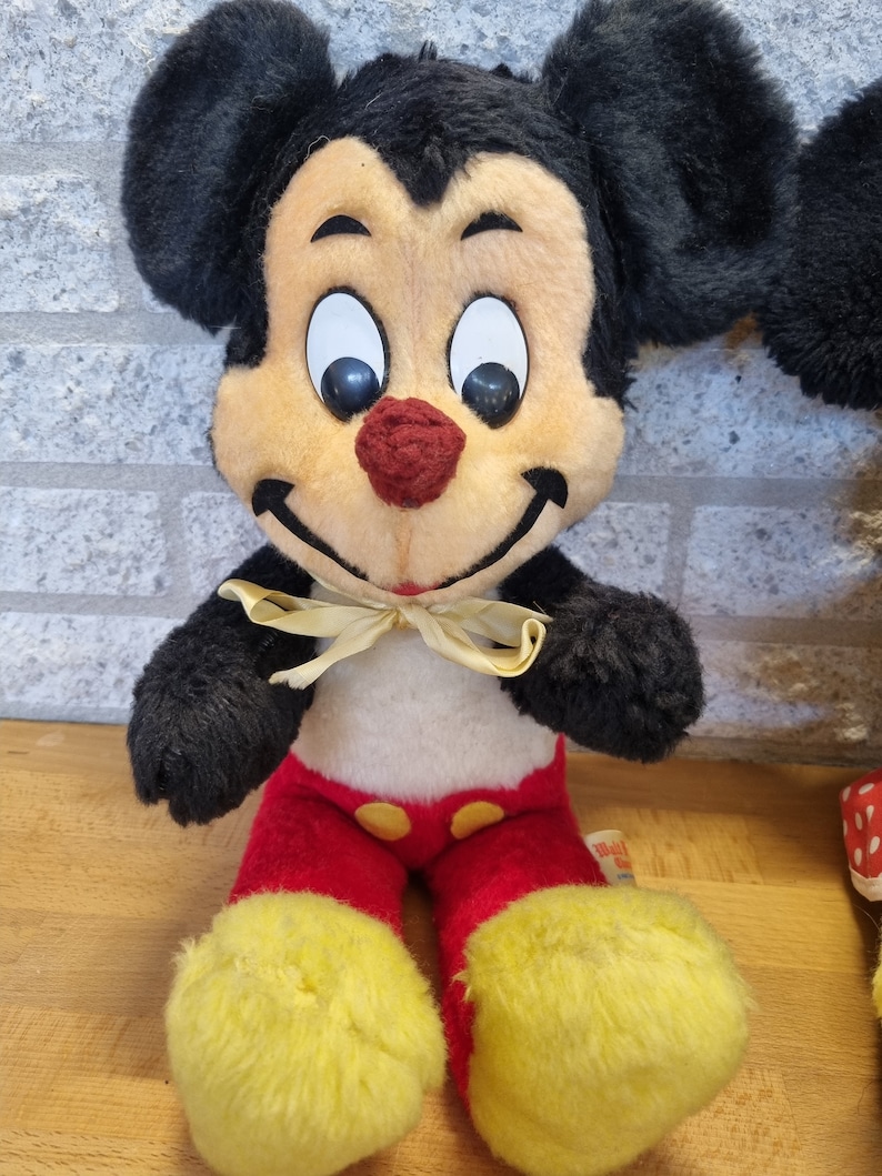May include: Vintage plush Mickey Mouse toy with black ears, a red and yellow outfit, and a yellow bow tie. The toy has a smiling face with black eyes and a red nose.