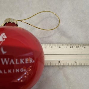 May include: A red glass Christmas ornament with the text "WALKER. WALKING" in white. The ornament has a gold-colored string for hanging. The ornament is approximately 3 inches in diameter.