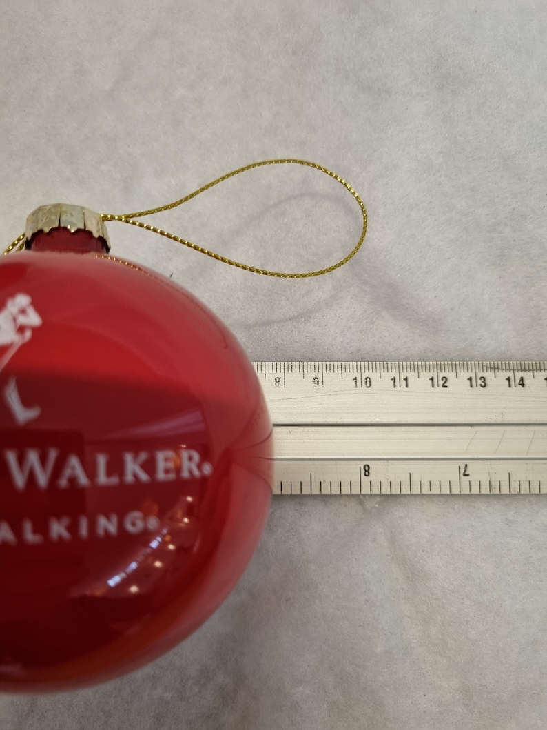 May include: A red glass Christmas ornament with the text "WALKER. WALKING" in white. The ornament has a gold-colored string for hanging. The ornament is approximately 3 inches in diameter.