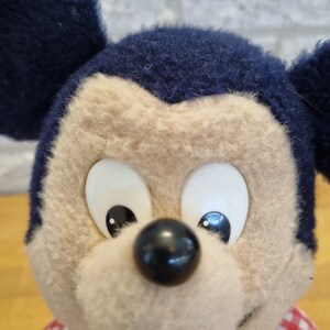 May include: Close-up of a plush Mickey Mouse doll. The doll has a tan face, large white eyes, and a black nose. It wears a blue denim overall and a red and white checkered shirt. The ears are dark blue.