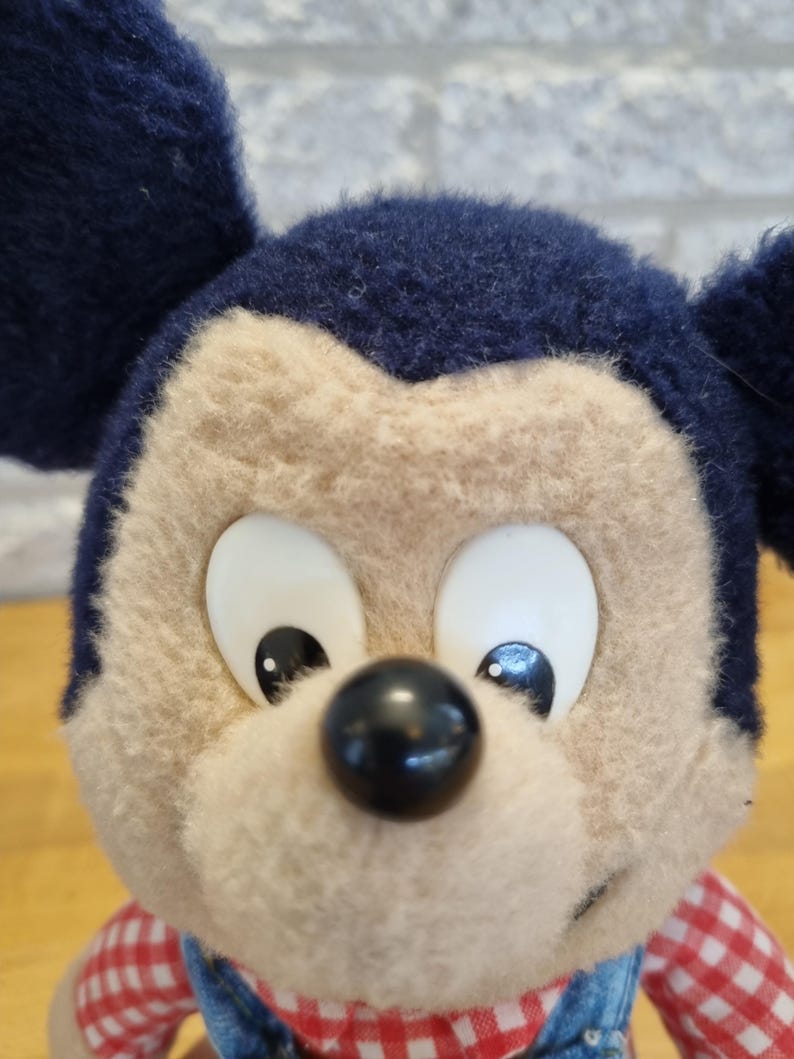 May include: Close-up of a plush Mickey Mouse doll. The doll has a tan face, large white eyes, and a black nose. It wears a blue denim overall and a red and white checkered shirt. The ears are dark blue.