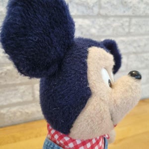 May include: A plush Mickey Mouse toy with navy blue ears, a tan face, and a black nose. He wears a red and white checkered shirt and denim overalls. The toy is soft and cuddly.